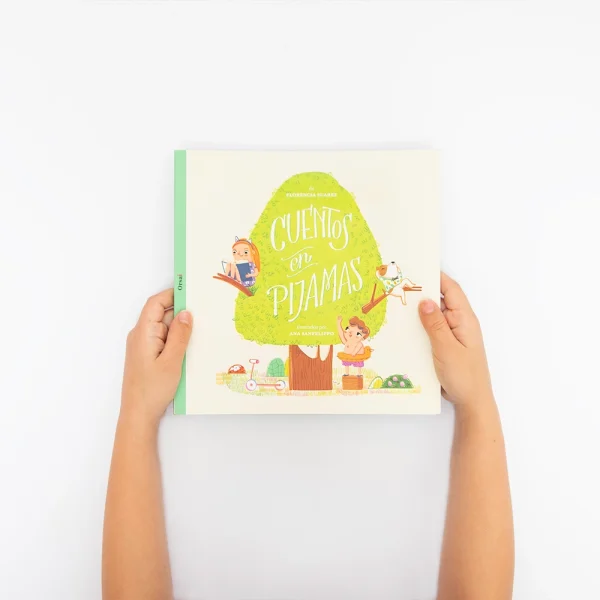 Dos manos infantiles sostienen el libro “Cuentos en Pijamas 1”. La portada muestra un árbol, una niña, un niño y un perrito.