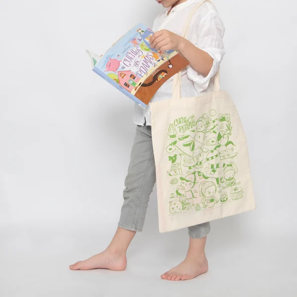 Niño sostendiendo un libro y una Totebag con ilustración en verde de Cuentos en Pijamas.