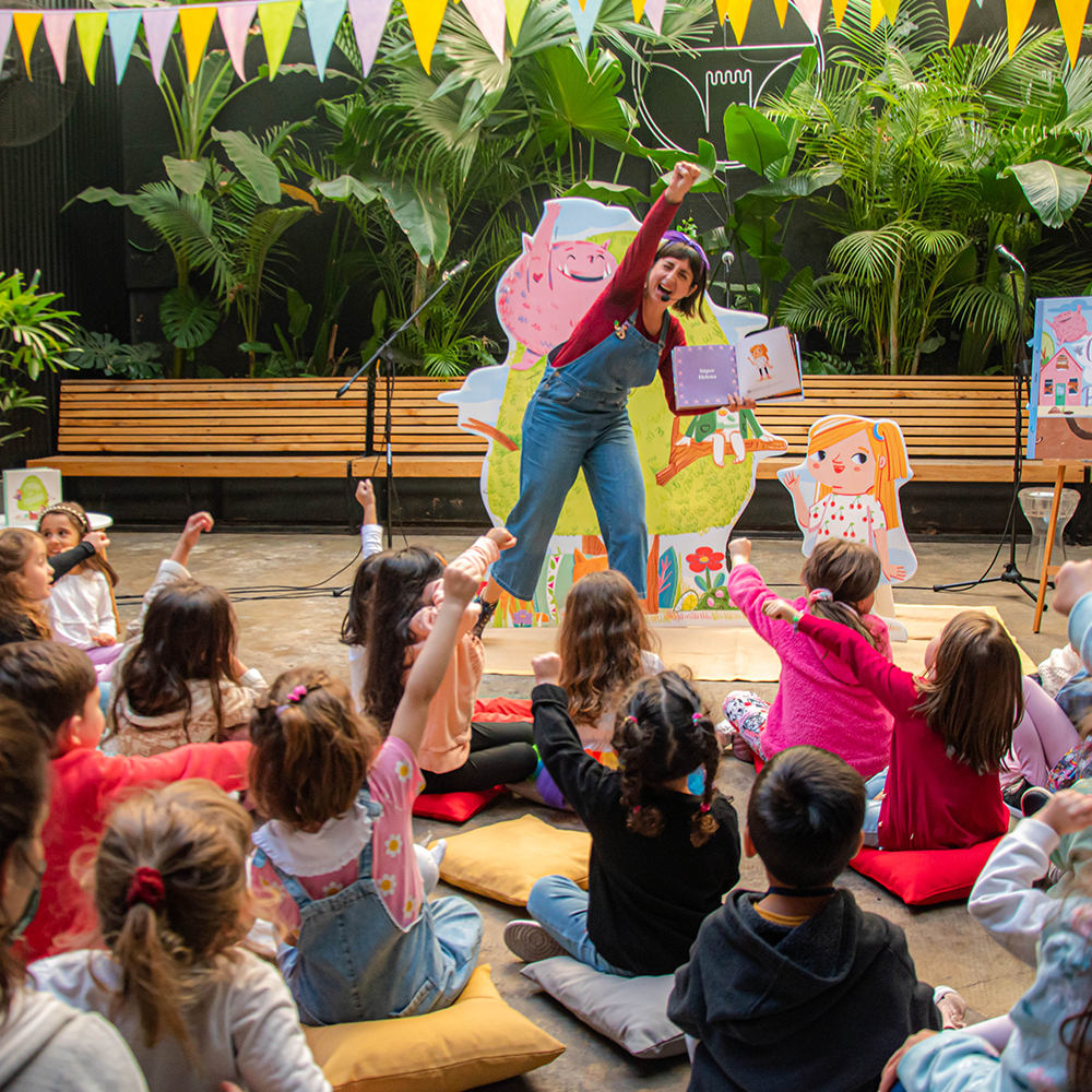 Fotografía de una cuentacuentos interactuando con un grupo de niños sentados en cojines, todos participando de manera entusiasta. La cuentacuentos, que sostiene un libro abierto y levanta el brazo en señal de entusiasmo, está rodeada de decoraciones coloridas y figuras de personajes ilustrados. El ambiente tiene plantas y guirnaldas de banderines, creando una atmósfera alegre y acogedora.